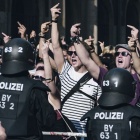 Wütende, brüllende Demonstranten recken den Mittelfinger in die Höhe. Vor der Menge, getrennt durch eine orange Absperrung stehen - mit dem Rücken zum Betrachter - behelmte Polizisten in schwarzer Schutzkleidung.