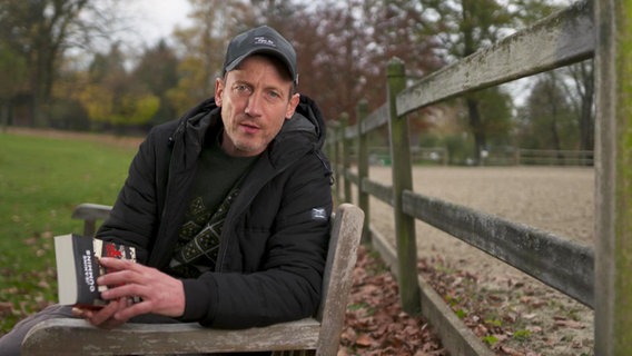 Wotan Wilke Möhring mit Buch auf einer Bank im Park.  