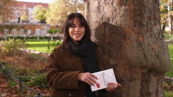 Almila Bagriacik mit Buch in den Händen neben einem Baum.  