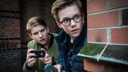 Johannes (Luke Matt Röntgen) hält ein Fotohandy und schaut mit Benny (Ruben Storck) um eine Hausecke. © NDR/Studio HH Fotograf: Boris Laewen