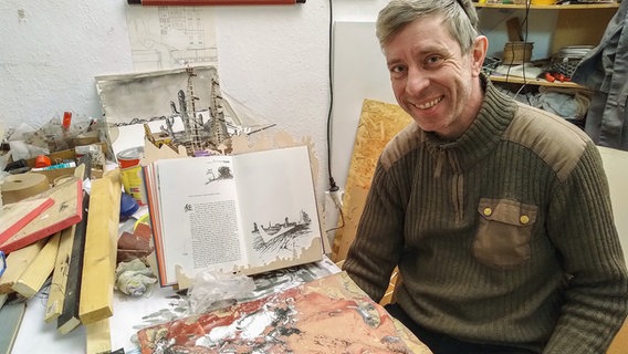 Der Illustrator und Künstler Henrik Schrat, ein Mann mit grauem Haar in mittleren Jahren, sitzt inmitten seiner Arbeitsutensilien. © NDR Foto: Franziska Storch