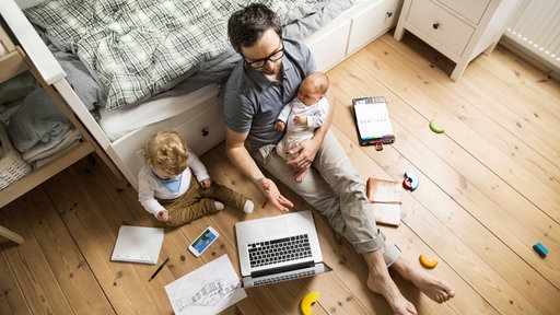 Ein Vater auf dem Fußboden in einem Schlafzimmer mit zwei Kindern und vor einem Laptop