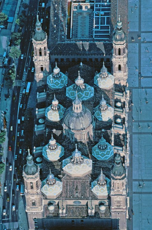 Saragossa, Hauptstadt der Provinz Aragonien, ist in Spanien ein ganz besonderer Ort. Spanien, das die Gründung seiner ersten christlichen Gemeinden. dem Apostel Jakobus zuschreibt, beansprucht für die Stadt Saragossa ausserdem eine weitere Auszeichnung: sie gilt als Schauplatz der ältesten Marienerscheinung. Dem am  Erfolg seiner Missionstätigkeit zweifelnden Apostel Jakobus erschien die Gottesmutter im Jahre 40, also noch zu Lebzeiten, ermunternd und tröstend auf einem Pfeiler aus Jaspis. In Saragossa lösten sich seither zu Ehren der Maria Pilar, der heiligen Jungfrau auf dem Pfeiler, immer neue Kapellen, Kirchen und andere Sanktuarien ab..Der Grundplan der jetzigen Wallfahrtskirche, ein rechteckiger Bau mit vier Ecktürmen und einer zentralen Kuppel stammt vpn 1681. Die mit farbigen glasierten Azulejo-Kacheln gedeckten Laternenkuppeln beleben die Dachlandschaft zusätzlich erst seit dem l9. Jahrhundert.

(Copyright Georg Gerster/Keystone)

www,georggerster.com