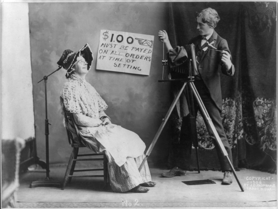 Der Porträtfotograf Das Foto enstand im Jahr 1902 irgendwo in Norddakota. Die Szenerie in einem improvisierten Fotostudio ist für heutige Augen eingermassen skurril. Das Schild gibt Auskunft darüber, dass für ein Foto ein Dollar zu entrichten ist, und zwar am Beginn der Sitzung für die Aufnahme.
Die damaligen Zeitgenossen haben allerdings über eine andere Pointe gelacht, die dem heutigen Betrachter zumindest auf den ersten Blick entgeht: Die ältere Dame ist in Wirklichkeit ein Mann. Wegen dieser Pointe hat die Library of Congress dieses Bild als humoristisch klassifiziert. (Foto: Library of Congress Prints and Photographs Division Washington/J. E. Pasonault)