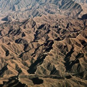  Die chinesische Mauer, unvorstellbar lang

Die „unvorstellbar lange Mauer“, so der chinesische Name des im 7. Jahrhundert v. Chr. begonnenen Bauwerks, ist nach der jüngsten amtlichen archäologischen Erhebung 21 136 km lang; die vormals meist genannte Zahl von 8 851 km bezog sich nur auf die in der Mingzeit erneuerten – und am besten erhaltenen – Teile der Mauer. Schätzungen, die alle Verästelungen, Seitenarme und Vorwerke berücksichtigen, kommen zum Schluss, dass sie sogar die Länge des Erdumfangs übertrifft. Legenden wollen wisssen, dass der König von Qin, der spätere Erste Erhabene Kaiser (221 bis 210 v,Chr.), im Traum auf dem Mond gelandet sei und von dort auf sein Reich zurückgeschaut habe. Es erschien ihm klitzeklein – so winzig, dass er beschloss, mit einer Mauer ein weit grösseres Gebiet als Vorgabe für sein zukünftiges Reich einzufrieden. Dieser Traum nährte vermutlich die Behauptung, die Chinesische Mauer sei das einzige vom Mond aus von blossem Auge erkennbare Menschenwerk. Ein Denkmal der Berührungsangst der Völker als einziges weithin sichtbares Signal für die Bewohnbarkeit dieses Planeten – wie traurig wäre das!. Aber Trost ist angesagt; die Behauptung ist nichts als nachgeplapperter Unsinn. Schon aus Satellitenflughöhe ist die Mauer schlechter erkennbar als etwa der Suezkanal oder die irdischen Ballungsräume, Peking eingeschlossen; und vom Mond aus überhaupt nicht. Der erste chinesische Raumfahrer, Yang Liwei, kehrte 2003 mit dem enttäuschenden Verdikt „nicht sichtbar“ zurück; der Staatsverlag änderte und druckte darauf einige Schulbücher neu. Die Doppelfunktion des Aus- und Einsperrens, die schon der kaiserliche Traum für die Mauer vorsah, erfüllte sie im jahrtausendelangen Auf und Ab der chinesischen Geschichte durchaus: sie hielt nicht nur die Barbaren fern, sondern begrenzte auch chinesische Expansion – jedenfalls zeitweilig. 1987 setzte die Unesco dieses stupende Sammelsurium von mehr als 40 000 Einzelobjekten und Standorten – Stücke von Wehrmauern und Schutzwällen, Wacht-, Leucht- und Signaltürme, Tore, Kasematten und Garnisonen, Arsenale und Exerzierplätze – auf die Liste des Welterbes. China erweitert seither laufend die touristische Zugänglichkeit der Mauer in der Nähe von Peking, des offenbar unvermeidlichen Vandalismus und des Mülls der Wegwerfgesellschaft wenig achtend. Das Flugbild zeigt die Mauer in den Bergen der Provinz Hebei bei Gubeikou. – Jahr der Aufnahme: 1987. (Copyright: Georg Gerster/Keystone)
