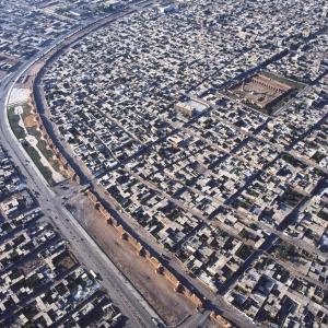 (Copyright: Georg Gerster/Keystone)

Die syrische Stadt ar-Raqqa am Euphrat ist seit 1999 Welterbe-Kandidat. Die radikal-sunnitische Terrormiliz „Islamischer Staat“ hat sie jetzt zur Hauptstadt ihres fantasierten Kalifats erkoren – ein Albtraum nicht nur für das Welterbekomitee. 
Eigentlich heisst ja die Stadt auf unserem Bild ar-Rafiqa („die Begleiterin“): der Abbasiden-Kalif al-Mansur gründete sie im Jahr 772 einige Steinwürfe weit von der hellenistisch-römisch-byzantinischen Stadt entfernt, die seit der arabischen Eroberung ar-Raqqa hiess. Von dem spätantiken ar-Raqqa gibt es nur noch Spuren. In der abbasidischen Schwesterstadt wirkt der kreisrunde, den Kosmos abbildende Grundriss Bagdads nach: der gewaltige Mauerring beschreibt einen hufeisenförmigen Bogen, dessen Öffnung vom Ufer des Euphrat geschlossen wird. Mit ihrer wachsenden Bedeutung an einem Kreuzweg wichtiger Handelsstrassen emanzipierte sich „die Begleiterin“ bald auch dem Namen nach. Jetzt wurde sie ar-Raqqa, während die spätantike Stadt Namen und Rang verlor. Kalif Harun ar-Raschid machte Raqqa von 796 bis 808 zu seiner Residenz, zwölf Jahre lang war es so das Verwaltungszentrum des abbasidischen Grossreichs.
Raqqa wurde als Festungsstadt an der Grenze zum byzantinischen Imperium konzipiert. Die Stadtmauer ragte über 18 Meter auf und ist mehr als 6 Meter mächtig. Backsteine ummanteln einen Kern aus ungebrannten Lehmziegeln. Etwa alle 25 Meter springen turmähnlich halbrunde Risalite zur flankierenden Bekämpfung von Angreifern vor. Von der urspünglich 4,5 Kilometer langen Mauer sind über 3 Kilometer erhalten, von den einst 132 Türmen stehen noch 75. Die Grosse Moschee („Freitagsmoschee“) im Stadtzentrum zeigt in der Bautechnik Ähnlichkeiten mit den Festungsbauten: ein mit Backsteinen verblenderter Kern aus Lehmziegeln. Den Grundplan der Anlage wie auch die ausgiebige Verwendung von Ziegeln statt von Hausteinen begreifen Architekturhistoriker als Nachhall aus mesopotamischer Zeit. Die Moschee ist jetzt Ruine, ihre Bethalle war ursprünglich überdacht.
Bei Ausbruch der syrischen Kriegswirren hatte Raqqa nahezu 300 000 Einwohner; im Flüchtlingsstrom seither ist es zu einer Millionenstadt angeschwollen. Dieser Stadtmoloch ist zurzeit ungemildert dem mörderischen Regime der radikal-sunnitischen Jihadisten „Islamischer Staat“ ausgeliefert, die in Raqqa die Hauptstadt ihres Kalifats sehen. Dem Vernehmen nach halten zwar die Besetzer die städtische Infrastruktur – Wasser- und Stromversorgung, Verkehr – einigermassen aufrecht, d.h. sie mischen sich bei den Betreibern nicht ein. Aber seit 2013 entehren und demolieren sie schiitische Moscheen, richten Andersgläubige hin, entweihen als eingeschworene Ikonoklasten christliche Kirchen, auferlegen den letzten in Raqqa ausharrenden Christen ein Kopfgeld, schliessen Schulen, zerstören die Grabmäler der Sufis auf Stadtgebiet. Und als zynischer Clou:  offenbar bedienen sie sich ungeniert in Raqqas Museen, der Verkauf von archäologischen Objekten auf dem Schwarzmarkt hilft ihren bilderstürmerischen Terror finanzieren. Raqqas kulturelle Substanz geht unwiederbringlich verloren, die einst angestrebte (und verdiente) Auszeichnung als Welterbestätte ist für immer verspielt. – Jahr des Flugbilds: 1997. (Copyright Georg Gerster/Keystone) 

