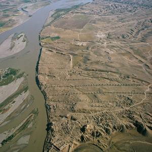 Assur am Tigris
Die irakischen assyrischen Königsstädte Nimrud und Ninive sind berühmter. Sie kommen ja auch in der Bibel vor, und erste Ausgrabungen dort um die Mitte des 19. Jahrhunderts lieferten monumentale Prachtsfunde. Dagegen spielte die Stadt Assur, eine Gründung des 3. vorchristlichen Jahrtausends, im 2.Jahrtausend durch Fernhandel reich und zur regionalen Macht geworden und vom 14.Jh. v. Ch. bis zum Anfang des 1.Jahrtausends die erste Hauptstadt des assyrischen Weltreichs, und darnach jahrhundertelang dessen Kulthauptstadt, die Rolle des Aschenbrödels. Das Alte Testament erwähnt sie mit keinem Wort, und frühe Grabungen, die ihre Ruinen anknabberten, erbrachten für Museen wenig Spektakuläres, die Schatzsucher wandten sich von dem spröden Objekt ab. Was ihm an Berühmtheit abging, machte es allerdings im 20. Jahrhundet an Ruhm wett. Im Auftrag der Deutschen Orientgesellschaft grub Walter Andrae in Assur von 1903 bis 1914. Die Grabung, die erste moderne Flächengrossgrabung mit dem vorrangigen Ziel der Informationsbeschaffung, und nicht  der Bereicherung europäischer Museen, schrieb Archäologiegeschichte. Bis heute gilt sie als methodisch mustergültig. Die Arbeitskräfte, fast 200 Arbeiter, hatte Andrae aus dem nahen Dorf Scherqat rekrutiert. Sie lernten soviel, dass sie, und später ihre Söhne und Enkel, während des ganzen Jahrhunderts auf allen Ausgrabungen im Irak bevorzugt beschäftigt wurden.„Scherqati“ heissen bis heute die Vorarbeiter auf Grabungen, sogar wenn sie gar nicht aus der Umgebung von Assur stammen. Andrae erschloss mit Sondiergräben das Stadtgelände. Die Wohnstadt interessierte den Bauforscher weniger als die Kernstadt mit ihren Tempeln, Palästen und Befestigungsmauern. Aquarellierend und zeichnend erweckte er  die Baureste zu dem Glanz der einstigen Götter- und Königsstadt. Im 9. Jh. v. Chr. verlegten die assyrischen Könige ihre Residenz nach Norden, zuerst wurde Nimrud, später Ninive das neue politische Machtzentrum. Assur freilich blieb die „heilige Stadt“ als Sitz des Reichsgottes, dem die Provinzgouverneure Schlacht- und Brandopfer schuldeten. Ein medisches Heer legte sie 614 v.Chr. in Schutt und Asche. Die Zeitläufte – zwei Weltkriege, die Teilung Deutschlands, zwei Irak-Kriege – behinderten die Rezeption der beispielhaften Grabung. Bis heute ist sie wissenschaftlich nicht vollständig aufgearbeitet. Seit den 1970er Jahren kümmerten sich irakische Archäologen immer mal wieder, restaurierend und grabend, um das Ruinengelände am Tigris. Zweimal kehrten auch deutsche Archäologen dorthin zurück, aber die Irak-Kriege machten stets einen Strich durch die Rechnung. Seit den 1980er Jahren gibt es Pläne für einen Damm, der den Tigris im Makhul-Gebirge (am oberen Bildrand) stauen soll. Der Stausee würde einen Teil des Stadtgeländes verschlingen. Die Unesco würdigte 2003 Asssur als Welterbe und setzte es gleichzeitig auf die rote Liste gefährdeter Denkmäler. Seither wurden zwar die Dammpläne schubladisiert – „endgültig“, heisst es in Baghdad, aber die Archäologen bleiben skeptisch. Zurzeit gilt das Dreieck Mosul-Kirkuk-Tigris mit Assur im Zentrum als so gefährlich, dass Feldforschungen oder auch nur Rekognoszierungen undenkbar sind. Auch deshalb leuchtet die Ampel der Unesco für Assur weiterhin warnend rot. Jahr der Aufnahme: 1973. (Copyright Georg Gerster/Keystone)