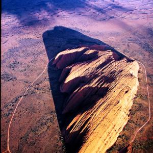 Der Uluru, 348 Meter über die umgebende Wüste emporragend, ist ein Berg der dritten Art: ganz eingesponnen in die „Traumzeit“ der örtlichen Ureinwohner.
Mag ja sein, dass der Fels im roten toten Herzen Australiens schon kraft seiner Natur weltweit einzigartig ist – mitnichten ist er aber ein Monolith, obwohl sogar die Unesco dieses Touristenlatein nachplappert. Und als Inselberg aus geschichtetem Sandstein, den Erosion und Verwitterung aus einer Rumpffläche herauspräparierten, ist er nicht einmal – so eine zweite ständig nachgebetete Behauptung – der grösste. Er ist ein Berg der dritten Art. Die Bezeichnung „heiliger Berg“ wird seiner Aura nicht gerecht, das Wort „heilig“ transportiert zu wenig Mythisch-Magisches. So berührungsscheu ist Uluru, dass die Gutachter in ihren Berichten zuhanden des Welterbekomitees auf gewisse Aspekte nur anspielen, sie aber keinesfalls ausführen durften. Sie zu bereden wäre soviel wie sie zu entweihen.

Dieser Berg hiess seit dem 19. Jahrhundert Ayers Rock (nach einem Ersten Sekretär von New South Wales und späterem australischen Premierminister) und wurde seit dem Bau einer Naturstrasse 1948 noch vor den Betonsegeln des Opernhauses in Sydney zum Markenzeichen Australiens. Die Naturikone war das Ziel einer lawinenartig anschwellenden Zahl von Schaulustigen (eingeschlossen das übliche Kontingent von Vandalen und Schmierern), die das Spektakel des bei Sonnenuntergang dramatisch erglühenden Bergs anzog. Den Ureinwohnern, die in Zentralaustralien seit wenigstens   30 000 Jahren nomadisieren und, nach Ausweis von Felsmalereien, mindestens seit 5 000 Jahren in Symbiose mit dem Berg leben, war dessen Herabwürdigung zum Tummel- und Rummelplatz ein Greuel. Australien schuf 1958 Abhilfe – auf fiese, zynische Weise: es nahm Ayers Rock und die nahen 36 Felsbuckel des Mount Olga (benannt nach einer Königin von Württemberg) den Ureinwohnern weg und erklärte sie zum Schutzgebiet; die Ureinwohner wurden nach Möglichkeit ausquartiert. 1977 avancierte das Schutzgebiet zum Nationalpark. Die Parkverwaltung beschnitt Auswüchse des Tourismusgewerbes, Hotels, Motels, Zelt- und Parkplätze mussten jetzt wenigstens die Parkgrenze respektieren. Die Ureinwohner freilich wollten ihren Berg zurück. Ihr Kampf endete nach zwischenzeitlichen Alibi-Ergebnissen (die Parkverwaltung stellte einige Ureinwohner als Rangers an) mit durchschlagendem Erfolg. 1985 ging der Rechtstitel am Parkland an die Ureinwohner über, die es im Gegenzug der Bundesregierung für 99 Jahre verpachteten. Gleichzeitig nahmen sie mit einer Mehrheit in dem Gremium Einsitz, das den Park verwaltet und betreibt.

Ayers Rock wurde Uluru, wie der Inselberg in der Sprache der ansässigen Pitjantjatjara – mit unklarer Bedeutung – heisst; und die Olgas wurden wieder Kata Tjuta („viele Köpfe“). Beim Namenswechsel blieb es nicht. Besucher mussten sich mit neuen Einschränkungen abfinden. Einige Stellen des Bergs sind stets tabu, und an Festtagen, an denen grosse Rituale die schöpferische „Traumzeit“ ins Jetzt und Hier holen, schliesst der Park. Die Ureinwohner sorgten aber für mehr als nur Verbote. Ihr in Jahrtausenden akkumuliertes ökologisches Wissen lommt dem Park zugute. Die Praxis kontrollierter Abbrände (für die Parkverwaltung bis anhin Anathema) erweist sich als Segen: das Feuer hilft Pflanzen mit essbaren Früchten keimen; auf den Brandflächen spriesst frisches Gras für Känguruhs. Einheimische Frauen säubern jährlich die natürlichen Zisternen, die am Fuss des Bergs das Regenwasser sammeln.

Der Park erlangte schon 1987 Welterbe-Würde; damals freilich noch, mit etwas wackliger Begründung, als Naturgut. Das Welterbekomitee doppelte l994 mit überzeugenderen Argumenten nach: nun würdigte es Uluru-Kata Tjuta als Kulturlandschaft und als unvergleichliche „assoziative Landschaft“. Das Wort „assoziativ“ ist (genauso wie „heilig“) bestenfalls ein Behelf. Uluru und Kata Tjuta deuten nicht auf die Heroen der „Traumzeit“ hin; sie sind Fels gewordene mythische Ahnen. Die Südseite von Uluru identifizieren die Anwohner mit den Python-Menschen und dem Blauzungenskink-Mann, die Nordwestecke mit der Beutelmulle-Frau. Trotzdem wurden bisher das Herumklettern auf dem Berg, seine Besteigung entlang einer Route über die Nordwestflanke nicht unterbunden. Die Parkverwaltung mahnt zwar die Touristen, nicht auf den Göttern der Ureinwohner herumzutrampeln und die Gipfeltour zu unterlassen, aber nicht gerade mit überwältigendem Erfolg. Ein beantragtes, klar formuliertes Besteigungsverbot ist unvermeidlich. – Jahr des Flugbilds: 1974 (Copyright Georg Gerster/Keystone)