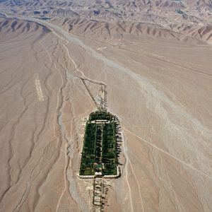 Persien schenkte uns nicht den Garten, aber es schenkte uns das Paradies, jedenfalls das Wort. Das altpersische Wort pairi daiza bezeichnete eigentlich nur einen ummauerten Raum, aber die Griechen nannten den Jagdgarten der persischen Könige paradeisos.

Die Unesco setzte 2011 neun Gärten in Iran auf die Liste des Welterbes. Sie sind über das ganze Land verstreut und gehören den verschiedensten Epochen an. Der älteste Garten, noch in Spuren erkennbar, gehörte zum Palastkomplex von Kyros dem Grossen (gestorben 530 v. Chr.) in Pasargadae. Der jüngste wurde von einem Kadscharenprinzen bei Mahan in der Provinz Kerman um die Mitte des 19. Jahrhunderts geplant und von dem Provinzgouverneur in den 1890er Jahren fertig gestellt : Bagh-e Shahzadeh, der „Prinzengarten“. (Er wird gelegentlich irrtümlich auch als Bagh-e Shah, „Garten es Königs“, bezeichnet.)  Die neun Gärten wandeln frei die Urform des persischen Gartens ab, der sich aus vier Quadranten zusammensetzt – eine symbolische Erinnerung an die zoroastrische Quaternität von Himmel, Erde, Wasser und Pflanzenwelt.

Der Prinzengarten ist ein längliches Rechteck mit einer Grundfläche von 5,5 ha. Von einer Eingangshalle führt ein zentraler Wasserlauf mit Becken und Kaskaden zwischen Zypressenalleen zu einem Schlösschen, das der Gouverneur als Sommerresidenz benützte. Dem Garten fliesst vom Gebirgsfuss her ober- und unterirdisch ganzjährig genügend Wasser zu. Die Wasserführung verzichtet auf Pumpen und verlässt sich ausschliesslich auf das Gefälle. Als Gartenschöpfung ist der Bagh-e Shahzadeh umstritten. Puristen, die auf die klassischen Gartenschöpfungen Persiens eingeschworen sind, rümpfen gerne die Nase. Sie nörgeln über die Pflanzordnung, die Pavillons, die Breite seiner Wasserläufe. Seine Lage hat freilich nicht ihresgleichen, das geben selbst Eiferer zu. Bei Mahan kommt der persische Garten, der nicht wie europäische Gärten sich in die Umgebung einfügt, sondern seiner Umgebung sperrig widerspricht, zu sich selber. Kahle, sengende, tote Wüste wird wie durch Magie in schattig-kühles Duften und Fruchten verwandelt – das Fegefeuer ins Paradies.

Nur als Postscriptum: solche Gartenparadiese wurden mitunter auch missbraucht, die Kühle fächelnder Palmen, das Säuseln der Bäume, das Duften der Blumen und Plätschern der Brunnen angereichert mit Drogen und weiblichem Liebreiz. DieAssassinen, eine Sekte, die zur Kreuzfahrerzeit den Gottesstaat wiederherstellen wollte, machten davon Gebrauch. Marco Polo berichtet von einem Assassinenoberen, der junge Männer, die für Selbstmordattentate rekrutiert worden waren, mit Opium betäubte und in eine Gartenanlage verlegte. Die Kontraktkiller, inmitten von Blumen und Jungfrauen zum Vernaschen aus ihrem Rausch erwacht, sollten einen realen Vorgeschmack auf das himmlische Paradies erhalten, das ihrer nach erfülltem Mordauftrag wartete. – Jahr der Aufnahme: 1977. (Copyright Georg Gerster/Keystone)