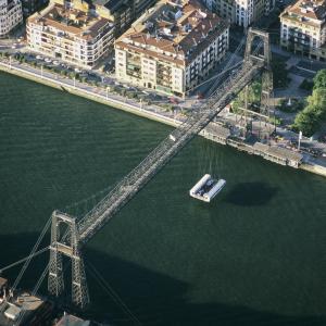 Die Schwebefähre über die Mündung des Flusses Nervión in den Golf von Biskaya ist die älteste der Welt.

Ihr Erfinder und Erbauer, der baskische Ingenieur und Architekt Alberto Palacio Elissague, verband 1893 die Orte Portugalete und Getxo im Ballungsraum von Bilbao, ohne den Schiffsverkehr in dem Ästuar zu behindern. Zwei 45 m hohe Stahlfachwerk-Türme stützen ein 160 m langes horizontales Traggerüst; an diesem hängt und fährt die Transportbarke. Die Biskaya-Brücke (baskisch Bizkaiko Zubia) ist noch heute im Betrieb. Sie setzt alle acht Minuten Menschen und Autos in anderthalb Minuten über und erspart ihnen zwanzig Kilometer Fahrt auf der Autobahn. Nachdem es einem jungen Lenker gelungen war, mitsamt Auto aus der Gondel tödlich abzustürzen, musste sich 2013 das Europäische Parlament mit einer Eingabe befassen, die unzureichende Sicherheitsvorkehrungen anmahnte.

Alberto Palacio war ein Schüler Gustave Eiffels, seine Schwebefähre in der Verknüpfung von Stahlbau mit Drahtseiltechnik zukunftweisend. An die zwanzig solcher Hochbrücken mit angehängter Transportbarke wurden in Europa, in beiden Amerikas und in Afrika gebaut. Acht von ihnen sind als Industriedenkmäler erhalten, einige noch betriebsbereit.

Die Schwebefähren haben weltweit eine rührige Fangemeinde. Seit zehn Jahren agiert ein Weltverband der Schwebefähren mit einer eigenen Webseite. In Deutschland haben sich um zwei Schwebefähren in Schleswig-Holstein (Rendsburg, Osten) spezielle Arbeitskreise gebildet. Alle Fans denken beim Stichwort Bilbao an die Biskaya-Brücke – und nicht etwa an das auf Blickfang angelegte Guggenheim–Museun. Die Auszeichnung der Biskaya-Brücke 2006 durch die Unesco als kulturelles Welterbe erfüllt sie, zugegeben, auch mit etwas Neid. Aber noch mehr mit Enthusiasmus. Sie bereiten zurzeit eine Sammel-Kandidatur für die restlichen Schwebefähren vor. – Jahr des Flugbilds: 2008 (Copyright Georg Gerster/Keystone)