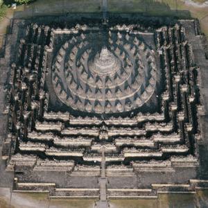 Der Borobudor auf der Indonesischen Insel Java
Der Borobudur ist weltweit ein Nonplusultra sakraler Baukunst: eine Stufenpyramide von 113 m Sockelbreite und 35 m Höhe mit fünf quadratischen und drei runden Terrassen, mit einem Bilderfries von 2,5 km Länge und über 400 Buddhafiguren. 1300 Reliefpaneele, die aus dem Leben der Bodhisattvas und des Buddha erzählen, verdeutlichen dem Pilger den Aufstieg aus der „Welt der Lüste“ durch „die Welt der Namen und Formen“ zu der erstrebten Läuterung. Der Stupa bildet das budhhistische Universum ab; Harmoniker hören ihn gar als in Stein gesetzte Musik. Schon der Bauherr, vermutlich ein König der Shailendras-Dynastie um 800, hatte freilich seine liebe Mühe mit der Fundamentierung; die untersten, die Welt des Karma illustrierenden Reliefpaneele verschwanden in einer sichernden Steinpackung, und die Silhouette des Tempelbergs wurde abgeflacht („une pâtisserie mal soufflée“ frotzelte später sakrilegisch ein Franzose). Der nahe Merapi, der auch wieder im Oktober 2010 obere Terrassen des Tempels zeitweilig mit saurer Vulkanasche unzugänglich machte, schneite ihn bei einem Grossausbruch im Jahr 1006 ein und der Dschungel frass ihn auf. Im 19. Jahrhundert zogen Briten die schützende Decke weg – und gaben ihn ungewollt dem Verderb durch Monsunregen, agresssive Algen, Flechten und Pilze preis. Eine radikale Sanierung 1975-1982 unter der Ägide der Unesco rettete das einsturzgefährdete Bauwerk in allerletzter Minute. Der Borobudur wurde völlig zerlegt und mit schweizerischer Klebetechnik und Araldit wieder zusammengeleimt. Die Mittel fehlten, um bei dieser Gelegenheit den verborgenen Fuss der Stufenpyramide zu befreien..Jahr der Aufnahme: 1989 (Copyright: Georg Gerster/Keystone)

 