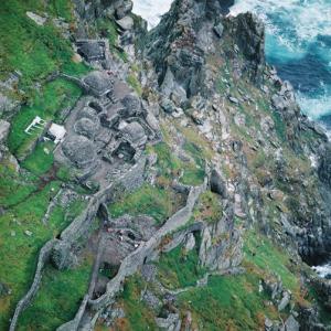 Skellig Michael, ein früher christlicher Vorposten am Rande der Welt, liegt vor Irlands Südwestküste. Was den Einsiedlern im Osten des damaligen christlichen Universums recht war, das war den Anachoreten im Westen billig: die Suche nach Abgeschiedenheit für ein Büsserleben im Gebet und in der Meditation. Jene zogen sich in die Wüste zurück und mal auch auf eine Säule; diese entsagten der Welt auf sturm- und wellengepeitschten Klippen vor der Atlantikküste.

Auf zwanzig Inselchen vor der Südwestküste Irlands allein finden sich Spuren früher Mönchsgemeinschaften; deren bauliches Erbe ist nirgends authentischer und besser erhalten als auf der Grossen Skellig. Was Wunder: es ist schwierig, einen abweisenderen, menschenfeindlicheren Standort als diesen zweigezackten, steilgeböschten Sandsteinfelsen 11,6 km vor der Iveragh-Halbinsel auch nur zu denken. Was hier Mönche und Einsiedler an Bausubstanz – fensterlose Zellen in Bienenkorbform und zwei bootsförmige Oratorien, alle in Trockenmauerwerk, dann eine Kirchenruine, Kreuze zwischen Gräbern – zurückliessen, das blieb in Gotteshand.

Der Insel wird jetzt auch ein vorgeschichtliches druidisches Observatorium angedichtet; das ist aber modisches Gemunkel, Druiden sind eben in. Die Geschichte der Insel begann erst in christlicher Zeit. Die Gottessucher waren todesmutig. Die Überfahrt in mit Leinen bespannten Booten aus Weidengeflecht gelang bestenfalls sommers, Winterwetter isolierte die Ankömmlinge für ein halbes Jahr. Trotzdem, oder gerade deswegen, liessen sich Büsser hier vielleicht schon im 6. Jahrhundert nieder.

Spätestens im 8. Jahrhundert gab es auf der Grosssen Skellig eine lose Mönchsgemeinschaft, anfangs wohl mehr Einsiedlerkolonie als eigentliches Kloster. Die Mitbewohner, viele Tausend Brutpaare von Papageitauchern, waren gut für Eier und Fleisch; das Meer lieferte Fische. Auf winzigen Anbauparzellen wuchsen Gemüse und Getreide. 823 hatten die Mönche ungebetenen Besuch: beutegierige Wikinger, erbost über die Habenichtse, verschleppten den Abt und liesssen ihn verhungern und verdursten. Eine dramatische Klimaverschlechterung vertrieb die Gottesleute im 12. Jahrhundert auf das milde irische „Festland“.

Dauerbewohner kamen erst wieder im 19. Jahrhundert: die Wärter eines Leuchtfeuers, das zuerst auf halber Höhe, dann, geschützter, weiter unten gebaut wurde. Geschützt? 1955 überspülte eine Flutwelle den immerhin 50 m über ruhigem Meer stehenden Leuchtturm, 1956 schwemmte die See einen Leuchtturmwärter weg. Jetzt ist das Leuchtfeuer automatisiert, der Turm unbemannt. Die Unesco. die   Skellig Michael 1996 Welterbe-Rang zuerkannte, begründete die Auszeichnung mit aussergewöhnlicher kultureller und architektonischer Zeugenschaft. Sie hätte auch ganz einfach an der Verzauberung eines sonst eher zum Spott aufgelegten (irischen) Besuchers Mass nehmen können: ein Ort mit „Magie, die einen hinaus, weit hinaus aus dieser Zeit und dieser Welt führt“, urteilte George Bernard Shaw. – Jahr des Flugbilds: 1989 (Copyright Georg Gerster/Keystone)