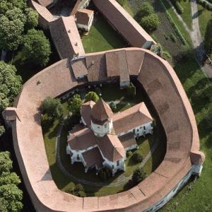 Von den erhaltenen 150 Kirchenburgen in Siebenbürgen – dem heutigen rumänischen Transsilvanien – setzte die Unesco sieben auf die Welterbe-Liste, ein denkmalpflegerischer Ansporn auch für den Restbestand. Tartlau, rumänisch Prejmer, eine von den sieben, ist als Welterbestätte ideal, zugleich musterhaft und einzigartig. Musterhaft für die Grundfunktion aller siebenbürgischen Kirchenburgen: Zuflucht für die (evangelische) Gemeinde in Notzeiten.mit vorbereiteter Infrastruktur: Schlafräume, Vorratskammern für Getreide und  Speckseiten, Mühle und Bäckerei, Zwinger für Schafe und Vieh  – gegebenenfalls für monatelange Belagerungen.

Tartlau ist die östlichste Gründung im vordem deutschen Siedlungsgebiet, seine Kirchenburg die eindruckvollste des Landes. Ihre graphische Eleganz – die Kreuzkirche eingeschrieben in einen runden Bering – macht sie einzigartig. Den Ort gründeten Deutschritter um 1212, der Bau der Kirche mit dem in Siebenbürgen singulären Grundriss eines griechischen Kreuzes begann wenig später. Befestigt wurde die Kirche erst im 15. und 16. Jahrhundert in laufender Anpassung an Neuerungen der Kriegstechnik. Der Bering – 14 m hoch, 5 m stark – erhielt einen Wehrgang mit Schiessscharten und Pechnasen sowie halbrunde Türme für die flankierende Verteidigung und wurde um eine Vorburg erweitert.

Die Innenseite der Ringmauer hat auf bis zu vier Stockwerken 250 Schlaf-, Kleider- und Vorratskammern. Keine Kirchenburg Siebenbürgens ist anziehender – und keine ist abweisender. Mit gutem Grund. Tartlaus Nähe zum Bosauer Pass, dem Einfallstor für die Reitervölker aus dem Osten, sorgte für besonders viele ungebetene Gäste: Mongolen, Türken, Tataren, Kosaken, Moldauer nebst assortierten Räubern, Freischärlern und Marodeuren. Der Ort wurde fast ein halbes hundert Mal gebrandschatzt und fast ebenso oft überlebten die Einwohner hinter den festen Mauern der Kirchenburg. – Jahr des Flugbilds: 1994  (Copyright Georg Gerster/Keystone)
