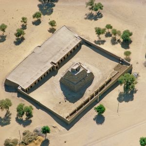 Das Reich der Malinke in Westafrika, das seit dem 13. Jahrhundert von der Atlantikküste bis nach Gao reichte. wurde im 15. Jahrhundert von dem Reich der Songhai abgelöst., das einen ähnlich grossen Teil des westsudanesischen Sahel belegte. Beide Königreiche wurden früh islamisiert, der Haddsch zweier Könige erregte Aufsehen sogar in Europa.

Vor allem der Mekkabesuch des Malikönigs Kankan Mansa Musa im Jahr 1325 gab zu reden. Er reiste mit grosssem Pomp, begleitet von 60&#039;000 Soldten. 500 Sklaven rannten voraus, jeder ausgerüstet mit einem zwei Kilogramm schweren Zeremonienstab aus purem Gold. Mit Gold warf der König nur so um sich, sodass das damalige Währungsgefüge ins Wanken geriet. Der Wert des ägyptischen Dinars halbierte sich. Mansa Musa selber wurde das Opfer dieser Entwertung. Bei der Rückkehr von Mekka war er knapp bei Kasse und lebte zeitweilig auf Pump.

Man müsste eigentlich annehmen, dass der Verschwender freigiebig war. Auf Almosen angewiesene Moscheen und Bedürftige erinnerten ihn aber eher als Geizknäpper. Hingegen Askia Mohammad Toure – wie anders! Auch seine Pilgerfahrt nach Mekka fast zwei Jahrhunderte später schimmerte golden. Der Herrscher des Songhaireichs, das die Kontrolle über den transsaharischen Handel mit Gold und Salz reich gemacht hatte, liess sich nicht lumpen. Aber er verteilte noch mehr Almosen und offenbar gezielter. Die Erinnerung an den frommen, in Übereinstimmung mit seinem Glauben mildtätigen Besucher lebte lange fort. Der Scherif von Mekka zeichnete ihn mit einem schwarzen Turban aus und ernannte ihn zum Imam. Der abbasidische Kalif von Kairo legalisierte seine Herrschaft und erlöste ihn von dem Stigma eines Usurpators.. Nach seiner Rückkehr aus Mekka machte er den Islam zur Staatsreligion. Als König kümmerte sich Mohammad besonders um die Armen, bei seinen Untertanen stand er in hohem Ansehen. Er schuf eine vorbildliche Verwaltung und führte einheitliche, verbindliche Masse und Gewichte sowie eine Währung ein. Er förderte die Wissenschaften und die Gelehrsamkeit, er hatte regen Umgang mit den heiligen Männern seines Reichs. Den Juden war freilich der Zutritt zum Songhai Reich verwehrt, aber der Kömig selber hielt sich nicht immer an das Verbot, das seinem liberalen Geist offenbar zuwider war.

Sein Grabmal plante er frühzeitig. Mehr als vierzig Jahre vor seinem Tod 1538, bei seiner Rückkehr aus Mekka im Jahr 1495 sollen 10&#039;000 Kamele Lehm und Holz für die Grabpyramide nach Gao, die Hauptstadt des Songhaireichs, gebracht haben. Dort entstand Askias letzte Ruhestätte mit zwei angebauten Moscheen, einem Friedhof und einem Versammlungsplatz – das grösste und höchste präkoloniale Bauwerk weitherum. Baumaterial war ungebrannter Lehm, der Baukörper ist gespickt mit vorstehenden Hölzern: dieses eingebaute Gerüst für Wartungsarbeiten gibt ihm ein igeliges Aussehen.

Askia der Grosse wurde in seiner Pyramide beigesetzt; sie war also kein Mausoleum. Folgekönige wurden in nächster Nähe bestattet. Die Unesco ehrte das Grabmal 2004 als Welterbe, musste es aber schon 2012 auf die rote Liste gefährdeter Welterbestätten setzen. Aufständische Tuareg und assortierte Jihadisten, die schon in Timbuktu ihr ikonoklastisches Mütchen gekühlt hatten, drohten auch dem Askia-Grab mit Unfug. – Jahr des Flugbilds: 1978 (Copyright Georg Gerster/Keystone)
