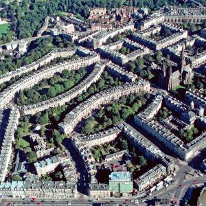 Schottlands Hauptstadt Edinburgh ist doppelgesichtig: neben der Altstadt „Auld Reekie“, der Alten Rauchigen, dehnt sich eine  elegante georgianische Neustadt aus.

Die alte Stadt besteht wesentlich aus einer einzigen Strasse, der „Königlichen Meile“. Sie verbindet die grimmige Burg am oberen Ende mit der Abtei und dem Palast von Holyroodhouse am unteren Ende eines Felskamms. Die Burg taucht au dem Dämmer der schottischen Frühgeschichte auf, den Bau des Palastes begann der Grossvater Maria Stuarts, Jakob IV., um 1500. In der Zeit englischer Invasionen oder bei Bruderzwisten zwischen König und der Nobilität in den Highlands befahl in Burg und Palast nicht notwendigerweise die gleiche Partei. Baugrund innerhalb der Stadtmauern war knapp. Die Hauptstrasse säumten vor allem Mietshäuser, sechs– bis zehngeschossig, damals so gut wie Wolkenkratzer, die Dächer gespickt mit Schornsteinen. Deren Russ und der Rauch der Kochstellen schwärzten die Mauern der Altstadt und verhalfen ihr unter den Edinburghern zu dem Kosenamen „Auld Reekie“. „Reeky“ mahnte in erster Linie die Verrussung und Verrauchung an, Kloakengeruch war allerdings immer mitgemeint. In diesem Stadtorganismus war die Königliche Meile das Rückgrat für Handel und Wandel; gleichzeitig fungierte sie freilich auch als sein Darm. Sie nahm Kot und Kehricht auf.

Die Realunion von l707 machte aus Schottland und England das Vereinigte Königreich Grossbritannien. Von nun an konnte das übervölkerte Edinburgh ungefährdet über die schützenden Stadtmauern hinauswachsen. Der Einstuz eines Mietshauses in der alten Stadt wirkte als Signal, der Plan einer neuen Stadt nördlich der alten nahm Gestalt an. Der Architekt James Craig, unbekannt und gerade mal 22 Jahre alt, gewann 1767 die Ausschreibung und machte sich unverzüglich ans Werk. Auf das Chaos der alten Stadt antwortete er im ersten Quartier der Neustadt mit einem strengen Schachbrettplan. Der Bau der New Town zog sich über ein volles Jahrhundert hin. Der westliche Teil um die Episkopalkirche St. Mary’s Cathedral – unser Bild – wurde erst in den 1880er Jahren 
fertig gebaut. Während dieser ganzen Zeit rührten die Architekten nicht an der pedantisch-eleganten Geometrie der klassizistischen Hausfassaden; die zusammengebauten Häuser kopieren fast zwanghaft das georgianische Muster. Dagegen traten die Urbanisten allmählich etwas lockerer auf. Sie nahmen Rücksicht auf das Gelände; die Häuserreihen bilden nicht mehr nur Quadrate und andere Vierecke, sondern auch Ellipsen, Halbmonde und Kreise. Man denkt natürlich an englische Städte, etwa an Bath; solche „crescents“ und „circuses“ im Stadtbild gehörten damals zum gängigen urbanistischen Vokabular. So löste ein stadtplanerischer Akt von hoher organisatorischer Disziplin das organische Wuchern der alten Stadt ab. In der Geschichte der europäischen Stadt belegt Edinburghs New Town ein Sonderkapitel. Die Auszeichnung als Welterbe im Jahr 1995 galt Edinburgh wohl vorwiegend als einem Beispiel georgianischer Stadtplanung, unübertroffen in Ausdehnung und Erhaltung. Die New Town war übrigens schon zu ihrer Zeit ein voller Erfolg. Magnetisch zogen die neuen Wohnquartiere die betuchten Edinburgher an. Es ist zu vermuten, dass diese nicht nur vor einstürzenden Häusern, Rauch und Russ Reissaus nahmen, sie hatten doch wohl schon lange die Nase voll, buchstäblich. Nach dem Auszug der bemittelten Bevölkerung vergammelte die Altstadt zeitweilig.

Nur nebenbei: auf sieben Hügeln erbaut, hätte Edinburgh den Vergleich mit Rom suchen können. Es wollte aber partout das Athen des Nordens sein. Ein Ehrenmal für die in den napoleonischen Kriegen gefallenen schottischen Soldaten und Matrosen sollte die Gestalt des Parthenons erhalten. Der ehrgeizige Bau begann 1822 auf dem Calton Hügel, aber die Finanzierung kam ins Stottern, der schottische Parthenon brachte es kläglich auf gerade zwölf Säulen. Das Fragment, jetzt als schottisches Nationaldenkmal im touristischen Angebot, erinnert zumindest an den Tod überbordender Griechenland-Begeisterung – und an die wirtschaftliche Realität des knappen Geldes, die ja den sprichwörtlich sparsamen Schotten ein Denkmal wert sein muss. – Aufnahmejahr des Flugbilds: 1999. (Copyright Georg Gerster/Keystone)