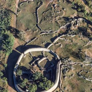 Pech für die im 19. Jahrhundert wiedergefundene Ruinenstadt im heutigen Staat Simbabwe: sie fiel gleich zu Anfang ihres zweiten Lebens unter die Phantasten.
Der schwäbische Geologe Karl Mauch hängte ihre Entdeckung 1871 an die grosse Glocke. Dem Kolumbus aus Stuttgart waren zwar einige Wikinger zuvorgekommen: Händler und Missionare hatten schon vor ihm von den überwucherten Steinbauten geraunt, Elefantenjäger neben ihnen kampiert – interesselos. Aber Mauch war überzeugt, das biblische Goldland Ophir gefunden zu haben, die Quelle von König Salomos Reichtum. Mehr noch: in den Bauten vermutete er die einstige zeitweilige Residenz Salomos und seiner Freundin, der Königin von Saba. Der Roman „King Solomon’s Mines“ (1886) von Henry Rider Haggard zementierte noch diesen Mumpitz. Der Bestseller brachte Sir Henry den Adelstitel und der Fundstelle einen zerstörerischen Goldrausch ein. Um 1900 waren für die nähere Umgebung 114 000 Claims eingetragen. Die Goldschürfer durchwühlten die Erde ohne Rücksicht auf archäologische Fragestellungen. Unter dem Mogeletikett „Ancient Ruins Company“ wütete mit behördlichem Segen eine Bande von Schatzjägern, die ihrem Namen Ehre machte. Sie liess wirklich nichts als Ruinen zurück. In einem Geschichtsbuch, das noch nicht einmal angelesen worden war, löschten sie Kapitel um Kapitel. Das 40 Hektar grosse Ruinengelände erstreckt sich zwischen zwei Baukomplexen. Auf einem Granithügel die festungsähnliche „Akropolis“ mit labyrinthisch verschachtelten Durchgängen und Räumen, verteidigungsfähig und als Sitz eines Potentaten denkbar. Und auf dem Talboden, zwischen Aloe und Wolfsmilch, eine monumentale Ellipse, die „Grosse Einfriedung“. Deren Umfassungsmauer, aus Granitsteinen mörtellos aufgeschichtet, 5 bis 11 m hoch, 1 – 5 m mächtig und 253 m lang, ist Afrikas grösster vorkolonialer Steinbau südlich der Sahara. Tempel? Zeremonielle Residenz des Königs? Ihre Funktion ist ungeklärt. Warum aber zum Verständnis der Ruinen Salomo und die Sabäerin bemühen? Die Weissen trauten den Schwarzen den Bau ganz einfach nicht zu. Dabei hatte die zweimalige archäologische Durchforstung, 1905 und 1929, alle Zweifel ausgeräumt: Gross-Simbabwe ist eine rein afrikanische Schöpfung, und zwar der Schona, eines Bantuvolks.

Gegründet bereits im 11.Jahrhundert, stieg es im 14. Jahrhundert zur Hauptstadt und zum Handelszentrum eines Staates auf, der von dem Goldreichtum der umliegenden Hochplateaus profitierte. Übervölkerung brachte Gross-Simbabwe schliesslich nach 1450 zu Fall: es mangelte an Nahrung und Feuerholz, die Schona mussten sich einen neuen Siedlungsplatz suchen. Die Portugiesen, die damals an der ostafrikanischen Küste aktiv wurden und dort die arabischen Kaufleute verdrängten, hörten von dem Goldumschlagplatz im Innern; schon sie flirteten mit der Idee des biblischen Ophir. Das Welterbekomitee nahm Gross-Simbabwe 1986 in die Familie der Welterbegüter auf, wohlwissend, dass es damit ein Sorgenkind mehr betreute. Verfahren zur nachhaltigen Konservierung von Trockenmauerwerk mussten erst ausgearbeitet werden. Zudem beeinträchtigt die touristische Erschliessung der Welterbestätte die archäologisch noch ungenügend explorierte weitere Umgebung. – Jahr des Flugbilds: 1983 (Copyright: Georg Gerster/Keystone).