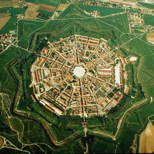  Nach der Juni-Tagung des Welterbekomitees im katarischen Doha gab es, aus den Bereichen Kultur und Natur, 1007 Welterbestätten. Ausserdem bewarben sich 1627 Kandidaten um die Aufnahme ins Pantheon.

Man darf ja auch einmal von Kandidaten sprechen. Mit 50 akzeptierten Welterbestätten (das halbe Hundert wurde am 22. Juni 2014 in Doha mit der Auszeichnung des piemontesischen Weinbaugebiets Langhe-Monferrato erreicht) steht Italien an der Spitze aller Vertragsstaaten, mit Vorsprung vor China, Spanien und andern Schwergewichten. Aber auch beim Wettstreit um das Welterbe kommt vielleicht der Appetit mit dem Essen. Im Falle Italiens, um bei dem Vorreiter zu bleiben, verzeichnet die Vorschlagsliste bereits 41 weitere Bewerbungen.

Zu den italienischen Kandidaten zählt Palmanova in Friaul-Julisch Venetien, ein Kleinod des Städtebaus. Palmanova ist eine jener Idealstädte, von denen in der Renaissance und im Barock viele entworfen, wenige gebaut wurden und noch weniger sich so lehrbuchmässig rein wie Palmanova erhalten haben. Das urbanistische Schema der Idealstadt ist vorwiegend rund und verrät so deren Abkunft von der „heiligen Stadt“ orientalischer Prägung. Ein viergeteilter Kreis versinnbildlichte die „heilige Stadt“, einen Mandala-ähnlichen Siedlungsarchetyp: den vom Horizont begrenzten Erdkreis mit den vier Weltgegenden. Freilich: die Abkunft der Idealstadt von der heiligen Stadt ist befleckt von dem Sündenfall ihrer Militarisierung, Idealstädte sind fast immer auch Festungsstädte.

Für die Serenissima Repubblica di San Marco, Venedig, war das Friaul ein Sorgenkind. Im ausgehenden 15. Jahrhundert hatten Türken siebenmal das Land verheert. Venedig beauftragte 1500 Leonardo da Vinci mit einem Gutachten zu den Verteidigungsanlagen im Friaul. Sie waren völlig ungenügend. Trotzdem entschloss sich Venedig erst 1593 zum Bau der Festungsstadt Palmanova; jetzt sah es sich nicht nur von den Osmanen, sondern auch von den Habsburgern bedroht. Die venezianischen Festungsingenieure sicherten die vom Reissbrett weg gebaute neue Stadt  mit einem ersten Verteidigungsring; er umfasste neun Bastionen zur flankierenden Bekämpfung von Angreifern und einen Graben. Im Laufe des 17. Jahrhunderts erhielt die Stadt einen zweiten Verteidigungsring. Und 1806, nach der Eroberung Palmanovas durch Napoleon, einen dritten. Die Strassen, Plätze und die Quartiere für Offiziere und Soldaten waren so angelegt, dass die Bollwerke im Blitz bemannt werden konnten. Der  allmähliche  Ausbau der Befestigungen echot die Fortschritte der Kriegstechnik, namentlich der Artillerie. Aber keine Erweiterung tastete das stadtplanerische Grundmuster des neunzackigen Sterns an.

Heutige Beschreibungen der Stadtgestalt heben stets den riesigen sechseckigen Platz im Mittelpunkt hervor; meist rügen sie ihn als überdimensioniert. Wurde er als Exerzierplatz der Garnison absichtlich übergross angelegt? Eine ganz andere Lösung für das Rätsel der Übergrösse fand ich im monumentalen Sechsbänder „Civitates Orbis Terrarum“, mit 363 Kupferstich-Veduten, erstmals publiziert 1572-1617 (jetzt als „Städte der Welt“ bei Taschen 2011). Der darin aufgenommene Stadtgrundnriss von Palmanova aus der Gründungszeit zeigt einen stark befestigten Wehrturm in der Mitte des Platzes – einen Donjon, einen Fluchtturm für Besatzung und Bewohner. Wurde der Turm schon bald nach der Stadtgründung abgerissen oder wurde er, sollte der Kupferstich lediglich eine Planskizze wiedergeben, gar nie gebaut? Wie dem auch sei, der Turm im Mittelpunkt passt akkurat zum Konzept der kosmischen Stadt. In deren orientalischer Ausführung gehörte der Mittelpunkt, der Nabel des Universums, dem Weltberg oder einem Tempelturm. Architektur ist ja immer auch Zeichensprache. Palmanova wäre also doch ein Abkömmling der heiligen Stadt. Auffällig ist ohnedies die Neunzahl der Bollwerke, der Bastionen, Ravelins, Kurtinen, Lünetten. Gleich dreimal die hochheilige Zahl 3, heiliger geht es wirklich nimmer.

Die Kandidatur Palmanovas wurde offiziell im Jahr 2006. Aber der Weg zum Welterbe ist weit und mühsam. Offenbar plagte das Städtchen die Angst, die mit der Anmeldung verbundene aufwendige Dokumentation nicht allein schultern zu können. Im Jahr 2011 suchte Palmanova Zuflucht in einem breiter angelegten, grenzüberschreitenden Welterbe-Vorschlag: „Die venezianischen Festungswerke zwischen dem 15. und l7. Jahundert“. Der Vorschlag bezieht ausser Palmanova die italienischen Städte Bergamo, Peschiera del Garda und Venedig sowie verwandte Festungsanlagen in Montenegro und Kroatien ein. Seit 2013 ist Palmanova auf Italiens Warteliste doppelt gemoppelt – Palmanova figuriert darauf als selbständiger Kandidat und gleichzeitig als Teil des Vorschlags „Venezianische Festungswerke“. Indes räumt die städtische Kulturreferentin in einem Telefongespräch ein, der Traum von einem Allleingang Palmanovas sei wohl ausgeträumt. – Jahr des Flugbilds: 1990 (Copyright Georg Gerster/Keystone)