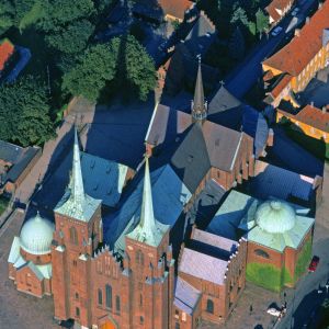 Copyright: Georg Gerster/Keystone
Meisterwerk der Backsteingotik

Die Kirche von Roskilde auf einem Geländebuckel am Ende des gleichnamigen Fjords ist der ältete Backsteindom Skandinaviens und die letzte Ruhestätte für 38 dänische Könige und Königinnen.

Die Kirche auf der dänischen Insel Seeland wurde 1280 nach mehr als hundert Jahren Bauzeit vollendet. Die Kathedrale hatte am selben Standort mehrere Vorgängerkirchen aus Holz oder Stein. Der Domkirche aus Backstein gaben die bischöflichen Bauherren, die in Paris studiert und dort zeitgenössische Tendenzen im französischen Kirchenbau verinnerlicht hatten, ein gotisches Gepräge. Sie gilt heute als eine frühe Vertreterin (und in ihrer Beispielhaftigkeit Wegbereiterin) der Backsteingotik in Nordeuropa. Von aussen wirkt die Kirche, ganz aus rostroten Ziegeln gebaut, wuchtig, aber wie  unerlöst. Erst wenn man sie betritt, wandelt sich Wuchtig in Erhaben. Jetzt stürmt die Kirche himmelwärts. Zu dem Eindruck erlöster Leichtigkeit in dem basilikalen Innenraum trägt viel der Umstand bei, dass weisser Verputz die ziegelroten Backsteinmauern zudeckt, aber Zier und Masswerk ausspart. Die ziegelroten Rippen, Stege, Rahmen und Einfassungen verleihen dem Dom die gotische Grafik..

Seit Anfang ist der rote Dom mit den grünspanigen Türmen und Kuppeln die Grablege des dänischen Königshauses. Die auch aus Vorgängerkirchen übernommenen Grabstätten reichen bis ins erste christliche Jahrtausend zurück. Die älteste ist diejenige des Wikingerkönigs Harald Blauzahn (910-987), der die erste Kirche an diesem Ort errichtete. Er war ein Reichseiniger - und sein Name lebt eben wegen seinem Genie des Verbindens in der elektronischen Welt von heute fort. Ein Ingenieur, der Wikingerromane verschlang, gab der Technologie kabelloser Datenübertragung zwischen zwei Geräten auf kurze Distanz den Namen Bluetooth; auch das Logo von Bluetooth, einer Kombination der Runen für H und B, bewahrt den Namen  König Blauzahns.

Die Kathedrale ist randvoll mit An- und Einbauten – Oratorien, Mausoleen und Grabkapellen. So voll, dass erstmals ein König, Frederik IX. (gestorben 1972), seit 1985 ausserhalb der Kirche in einem Backsteinbau beigesetzt ist. Die Grabkapellen im Innern des Doms dagegen schwelgen in Marmor (zum Teil  grönländischer Herkunft), die Sarkophage sind aus Granit, aus Alabaster, aus Marmor und Basalt (von den Färöern). Architektur und Schmuck der Kapellen spiegeln den Zeitgeist und die Vorlieben des Grabherrn. Christian I. (gestorben 1481) liess seine Grabkapelle mit Wandmalereien schmücken, wurde aber mit seiner Königin unter dem Boden der Kapelle bestattet. Christian III. (gestorben 1559) und Frederik II. (gestorben 1588) bezogen später die gleiche Kapelle; ihre Renaissance-Gräber gleichen miniaturisierten antiken Tempeln. In dieser Dreikönigskapelle (benannt nach den auf einem Fresko dargestellten drei Weisen aus dem Morgenland) steht Roskildes Kuriosum: die „Königssäule“. Königliche Besucher liessen darauf ihre Körpergrösse registrieren. Der Grabherr Christian I. führt die Liste mit 219, 5 cm an – fast 10 cm mehr als der nächstfolgende Herrscher, Zar Peter der Grosse. Freilich, Christians Übermass kam entweder durch Schlamperei beim Messen oder durch königlichen Schummel zustande – in Wahrheit war Christian knapp 189 cm gross. Immerhin war er also,  in nachmaliger preussischer Nomenklatur, ein Langer Kerl mit Gardemass.

In der Grablege der Könige suchten auch andere ihre letzte Ruhe; Hunderte kirchliche und weltliche Notablen, die es sich leisten konnten, sind in der Kathedrale beigesetzt, meist unter dem Boden des Kirchenschiffs. Der Boden ist eine einzige Sammlung von reliefierten Grabplatten. Sie mit Absperrungen zu schützen, ist unmöglich, die Mobilität in der Kirche würde unterbunden. So stapfen und stiefeln, trampeln und trippeln denn in diesem steinernen Friedhof die Pilger und Touristen über die Gräber und Toten. Wo bleibt da die Totenruhe? Oder auch nur die denkmalpflegerische Besorgnis: ist nicht der Abrieb der Reliefs auf den Grabplatten durch das Schuhwerk der Besucher zu befürchten? Die Unesco, die den Dom zu Roskilde 1995 auf die Liste der Welterbestätten setzte, über deren Schutz sie pingelig wacht, hatte keine Einwände. Und der diensttuende Kustos zerstreute bei einem Besuch vor einigen Wochen meine Bedenken. „Diese Grabplatten haben Pilgerfüsse, barfuss oder beschuht, tausend Jahre lang schadlos ausgehalten – und sie werden in nochmals tausend Jahren keinen Schaden nehmen.“ Die Nekropole Roskilde ist offenbar auch ein Ort der Hoffnung und Zuversicht... Jahr des Flugbilds: 1971. (Copyright Georg Gerster/Keystone)



