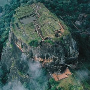 Sigiriya - König Kassapas Löwenfels
Jäh steigt Sigiriya im Herzen Sri Lankas 180 Meter aus dem umliegenden Dschungel empor: der Inselberg ist die Schlotfüllung eines wegerodierten Vulkans. Der singhalesische König Kassapa (473 – 491) baute auf diesem Magmapfropfen seine befestigte Residenz und an seinem Fuss eine neue Haupstadt.

Anuradhapura war ihm zu unsicher geworden, nachdem er dort seinen Vater hatte ermorden lassen. Er benützte den Berg nicht nur als Gründung für Aufbauten, sondern gestaltete ihn zur Architekturplastik um. Sogar die Felsblöcke, die unverrückbar am Fuss des Steilfelsens liegen, bezogen seine Gestalter in die Gesamtanlage ein. Diese können wir heute freilich nur noch erahnen. Kassapa schwebte anscheinend eine Art Götter- und Himmelsberg vor. Dessen Westseite liess er mit Putz bewerfen und vielleicht mit lauter Bildern von Blumen opfernden, auf Wolken thronenden Nymphen bemalen. Zweifellos sind die erhaltenen Fresken lediglich ein bescheidener Rest des ursprünglichen Schmucks, der im Schutze eines natürlichen Felsdachs Witterung und Vandalen (und Restauratoren) überlebte.

Lebte der Usurpator Kassapa auf dem uneinnehmbaren Felsen in Angst vor seinem Halbbruder, dem rechtmässigen Thronerben? Zwar umgürteten Wall und Graben die Stadt am Fuss des Burgfelsens. Aber Sigiriya macht insgesaamt eher einen heiteren Eindruck. Vorab die noch heute erkennbare planerische Umsicht, die dieses Versailles im Urwald schuf, sicherte Sigiriya 1982 einen Platz auf der Liste des Welterbes. Ausgedehnte Kunstgärten sind dem Felsen vorgelagert, mit Teichen, Schwimmbecken und Wasserspielen. Es fehlten auch nicht Badehäuser und Pavillons.

Der Anstieg führt durch den Schuttfuss des Bergs mit zyklopischen Felsblöcken, die Kassapas Steinmetzen, Maurer und Zimmerleute in Empfangsräume, Thronsäle und Heiligtümer verwandelt hatten. Eine Galerie, die ein natürliches Felssims ausnützt, nimmt zuletzt den Besucher auf. Ihre gemauerte Brüstung ist innenseitig mit einem glänzenden, polierten Bewurf versehen. Entlang dieser Spiegelwand, die noch teilweise erhalten ist, ging man in Kassapas Zeiten um den halben Berg herum, bis auf den Absatz mit dem kolossalen Löwen, der dem Berg den Namen Sigiriya, das heisst „Löwenfels“, gab.Von dem Riesentier aus Ziegeln und Stuck sind noch die Vorderpfoten zu sehen. Die Treppe, die den Absatz und die Palastanlage auf dem terrassierten Gipfel verbindet, muss anfangs im Innern des Tiers verlaufen sein. Die Reste auf dem Gipfelplateau deuten ebenfalls auf das süsse Leben des Vatermörders hin. Keine Spur von einer finsteren Fluchtburg. Dafür, ausser vielfältigen Anlagen zum Einfangen und Aufbewahren des Regenwassers, Swimmingpools, Lustgemächer, ein Hochsitz mit Thronhimmel...Was wunder, wenn die örtlichen Fremdenführer die gemalten Blumenmädchen gerne als Pin-ups von Kassapas Garnison deuten. Das ist natürlich der bare Unsinn. Doch gedeiht er, weil sich die Forschung in der Deutung der Malereien selber uneins ist. Mit dem guten Leben war es nach achtzehn Jahren vorbei.

Der um den Thron betrogene Halbbruder kehrte mit einer Armee aus dem indischen Exil zurück, Kassapa gab sich nach einer verlorenen Schlacht selber den Tod, Regierungssitz wurde wieder Anuradhapura. Menschenrechtwächter ermahnen heute Sri Lanka-Reisende, vor den Menschenrechtsverletzungen, die seit dem Ende des Bürgerkriegs weiterhin an der Tagesordnung sind, nicht die Augen zu verschliessen. Der Vatermörder Kassapa und assortierte Schurken aus dem Mahawamsa, der Grossen Chronik, sollen das Empörungspotential der Besucher nicht völlig aufbrauchen.– Jahr des Flugbilds: 1969. (Copyright Georg Gerster/Keystone)