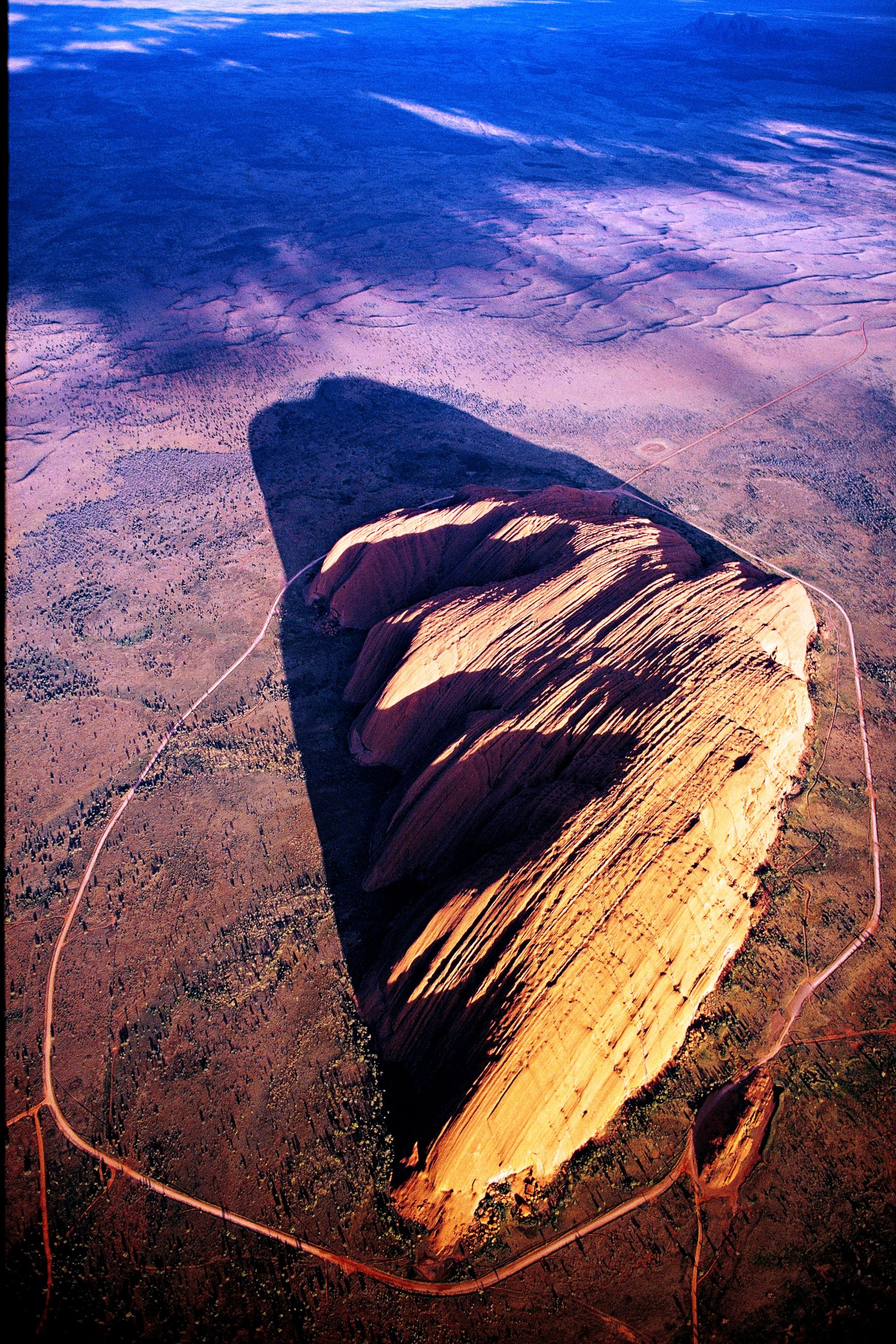 Der Uluru, 348 Meter über die umgebende Wüste emporragend, ist ein Berg der dritten Art: ganz eingesponnen in die „Traumzeit“ der örtlichen Ureinwohner.
Mag ja sein, dass der Fels im roten toten Herzen Australiens schon kraft seiner Natur weltweit einzigartig ist – mitnichten ist er aber ein Monolith, obwohl sogar die Unesco dieses Touristenlatein nachplappert. Und als Inselberg aus geschichtetem Sandstein, den Erosion und Verwitterung aus einer Rumpffläche herauspräparierten, ist er nicht einmal – so eine zweite ständig nachgebetete Behauptung – der grösste. Er ist ein Berg der dritten Art. Die Bezeichnung „heiliger Berg“ wird seiner Aura nicht gerecht, das Wort „heilig“ transportiert zu wenig Mythisch-Magisches. So berührungsscheu ist Uluru, dass die Gutachter in ihren Berichten zuhanden des Welterbekomitees auf gewisse Aspekte nur anspielen, sie aber keinesfalls ausführen durften. Sie zu bereden wäre soviel wie sie zu entweihen.

Dieser Berg hiess seit dem 19. Jahrhundert Ayers Rock (nach einem Ersten Sekretär von New South Wales und späterem australischen Premierminister) und wurde seit dem Bau einer Naturstrasse 1948 noch vor den Betonsegeln des Opernhauses in Sydney zum Markenzeichen Australiens. Die Naturikone war das Ziel einer lawinenartig anschwellenden Zahl von Schaulustigen (eingeschlossen das übliche Kontingent von Vandalen und Schmierern), die das Spektakel des bei Sonnenuntergang dramatisch erglühenden Bergs anzog. Den Ureinwohnern, die in Zentralaustralien seit wenigstens   30 000 Jahren nomadisieren und, nach Ausweis von Felsmalereien, mindestens seit 5 000 Jahren in Symbiose mit dem Berg leben, war dessen Herabwürdigung zum Tummel- und Rummelplatz ein Greuel. Australien schuf 1958 Abhilfe – auf fiese, zynische Weise: es nahm Ayers Rock und die nahen 36 Felsbuckel des Mount Olga (benannt nach einer Königin von Württemberg) den Ureinwohnern weg und erklärte sie zum Schutzgebiet; die Ureinwohner wurden nach Möglichkeit ausquartiert. 1977 avancierte das Schutzgebiet zum Nationalpark. Die Parkverwaltung beschnitt Auswüchse des Tourismusgewerbes, Hotels, Motels, Zelt- und Parkplätze mussten jetzt wenigstens die Parkgrenze respektieren. Die Ureinwohner freilich wollten ihren Berg zurück. Ihr Kampf endete nach zwischenzeitlichen Alibi-Ergebnissen (die Parkverwaltung stellte einige Ureinwohner als Rangers an) mit durchschlagendem Erfolg. 1985 ging der Rechtstitel am Parkland an die Ureinwohner über, die es im Gegenzug der Bundesregierung für 99 Jahre verpachteten. Gleichzeitig nahmen sie mit einer Mehrheit in dem Gremium Einsitz, das den Park verwaltet und betreibt.

Ayers Rock wurde Uluru, wie der Inselberg in der Sprache der ansässigen Pitjantjatjara – mit unklarer Bedeutung – heisst; und die Olgas wurden wieder Kata Tjuta („viele Köpfe“). Beim Namenswechsel blieb es nicht. Besucher mussten sich mit neuen Einschränkungen abfinden. Einige Stellen des Bergs sind stets tabu, und an Festtagen, an denen grosse Rituale die schöpferische „Traumzeit“ ins Jetzt und Hier holen, schliesst der Park. Die Ureinwohner sorgten aber für mehr als nur Verbote. Ihr in Jahrtausenden akkumuliertes ökologisches Wissen lommt dem Park zugute. Die Praxis kontrollierter Abbrände (für die Parkverwaltung bis anhin Anathema) erweist sich als Segen: das Feuer hilft Pflanzen mit essbaren Früchten keimen; auf den Brandflächen spriesst frisches Gras für Känguruhs. Einheimische Frauen säubern jährlich die natürlichen Zisternen, die am Fuss des Bergs das Regenwasser sammeln.

Der Park erlangte schon 1987 Welterbe-Würde; damals freilich noch, mit etwas wackliger Begründung, als Naturgut. Das Welterbekomitee doppelte l994 mit überzeugenderen Argumenten nach: nun würdigte es Uluru-Kata Tjuta als Kulturlandschaft und als unvergleichliche „assoziative Landschaft“. Das Wort „assoziativ“ ist (genauso wie „heilig“) bestenfalls ein Behelf. Uluru und Kata Tjuta deuten nicht auf die Heroen der „Traumzeit“ hin; sie sind Fels gewordene mythische Ahnen. Die Südseite von Uluru identifizieren die Anwohner mit den Python-Menschen und dem Blauzungenskink-Mann, die Nordwestecke mit der Beutelmulle-Frau. Trotzdem wurden bisher das Herumklettern auf dem Berg, seine Besteigung entlang einer Route über die Nordwestflanke nicht unterbunden. Die Parkverwaltung mahnt zwar die Touristen, nicht auf den Göttern der Ureinwohner herumzutrampeln und die Gipfeltour zu unterlassen, aber nicht gerade mit überwältigendem Erfolg. Ein beantragtes, klar formuliertes Besteigungsverbot ist unvermeidlich. – Jahr des Flugbilds: 1974 (Copyright Georg Gerster/Keystone)
