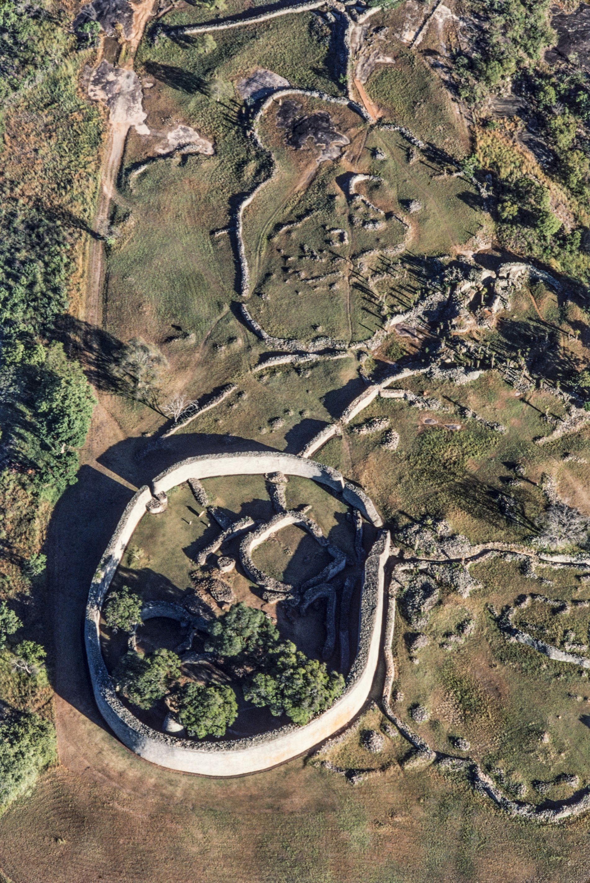 Pech für die im 19. Jahrhundert wiedergefundene Ruinenstadt im heutigen Staat Simbabwe: sie fiel gleich zu Anfang ihres zweiten Lebens unter die Phantasten.
Der schwäbische Geologe Karl Mauch hängte ihre Entdeckung 1871 an die grosse Glocke. Dem Kolumbus aus Stuttgart waren zwar einige Wikinger zuvorgekommen: Händler und Missionare hatten schon vor ihm von den überwucherten Steinbauten geraunt, Elefantenjäger neben ihnen kampiert – interesselos. Aber Mauch war überzeugt, das biblische Goldland Ophir gefunden zu haben, die Quelle von König Salomos Reichtum. Mehr noch: in den Bauten vermutete er die einstige zeitweilige Residenz Salomos und seiner Freundin, der Königin von Saba. Der Roman „King Solomon’s Mines“ (1886) von Henry Rider Haggard zementierte noch diesen Mumpitz. Der Bestseller brachte Sir Henry den Adelstitel und der Fundstelle einen zerstörerischen Goldrausch ein. Um 1900 waren für die nähere Umgebung 114 000 Claims eingetragen. Die Goldschürfer durchwühlten die Erde ohne Rücksicht auf archäologische Fragestellungen. Unter dem Mogeletikett „Ancient Ruins Company“ wütete mit behördlichem Segen eine Bande von Schatzjägern, die ihrem Namen Ehre machte. Sie liess wirklich nichts als Ruinen zurück. In einem Geschichtsbuch, das noch nicht einmal angelesen worden war, löschten sie Kapitel um Kapitel. Das 40 Hektar grosse Ruinengelände erstreckt sich zwischen zwei Baukomplexen. Auf einem Granithügel die festungsähnliche „Akropolis“ mit labyrinthisch verschachtelten Durchgängen und Räumen, verteidigungsfähig und als Sitz eines Potentaten denkbar. Und auf dem Talboden, zwischen Aloe und Wolfsmilch, eine monumentale Ellipse, die „Grosse Einfriedung“. Deren Umfassungsmauer, aus Granitsteinen mörtellos aufgeschichtet, 5 bis 11 m hoch, 1 – 5 m mächtig und 253 m lang, ist Afrikas grösster vorkolonialer Steinbau südlich der Sahara. Tempel? Zeremonielle Residenz des Königs? Ihre Funktion ist ungeklärt. Warum aber zum Verständnis der Ruinen Salomo und die Sabäerin bemühen? Die Weissen trauten den Schwarzen den Bau ganz einfach nicht zu. Dabei hatte die zweimalige archäologische Durchforstung, 1905 und 1929, alle Zweifel ausgeräumt: Gross-Simbabwe ist eine rein afrikanische Schöpfung, und zwar der Schona, eines Bantuvolks.

Gegründet bereits im 11.Jahrhundert, stieg es im 14. Jahrhundert zur Hauptstadt und zum Handelszentrum eines Staates auf, der von dem Goldreichtum der umliegenden Hochplateaus profitierte. Übervölkerung brachte Gross-Simbabwe schliesslich nach 1450 zu Fall: es mangelte an Nahrung und Feuerholz, die Schona mussten sich einen neuen Siedlungsplatz suchen. Die Portugiesen, die damals an der ostafrikanischen Küste aktiv wurden und dort die arabischen Kaufleute verdrängten, hörten von dem Goldumschlagplatz im Innern; schon sie flirteten mit der Idee des biblischen Ophir. Das Welterbekomitee nahm Gross-Simbabwe 1986 in die Familie der Welterbegüter auf, wohlwissend, dass es damit ein Sorgenkind mehr betreute. Verfahren zur nachhaltigen Konservierung von Trockenmauerwerk mussten erst ausgearbeitet werden. Zudem beeinträchtigt die touristische Erschliessung der Welterbestätte die archäologisch noch ungenügend explorierte weitere Umgebung. – Jahr des Flugbilds: 1983 (Copyright: Georg Gerster/Keystone).